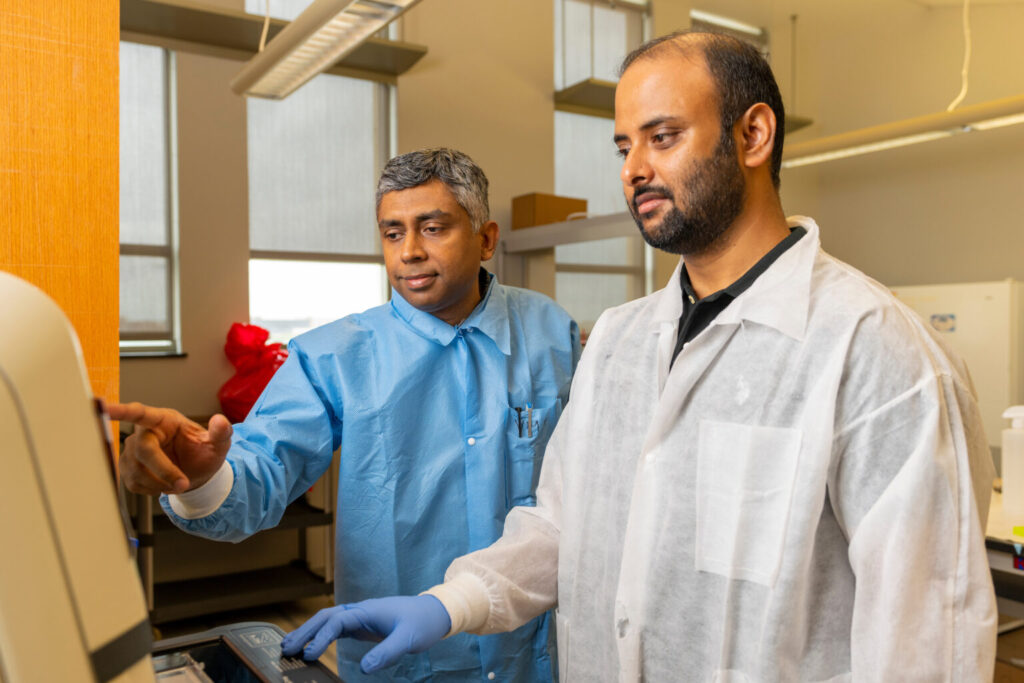 Two men, Vishal Gohil and Mohammad Zulkifli stand in a biology lab examining a Petri dish that Gohil is holding. The man on the left is wearing a blue lab coat and the one of the right is wearing a white lab coat. 