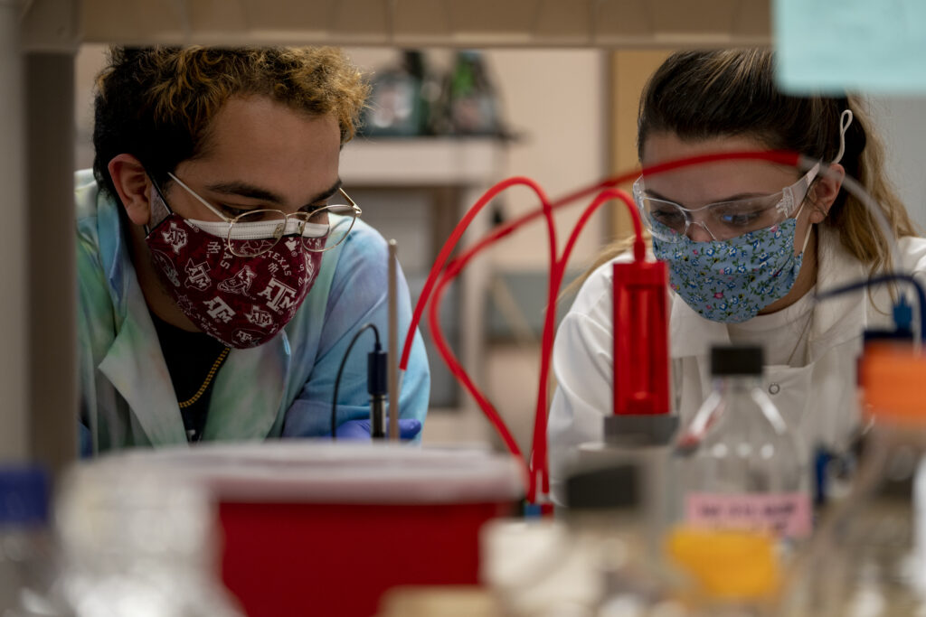 Two students wearing masks and lab coats are in lab