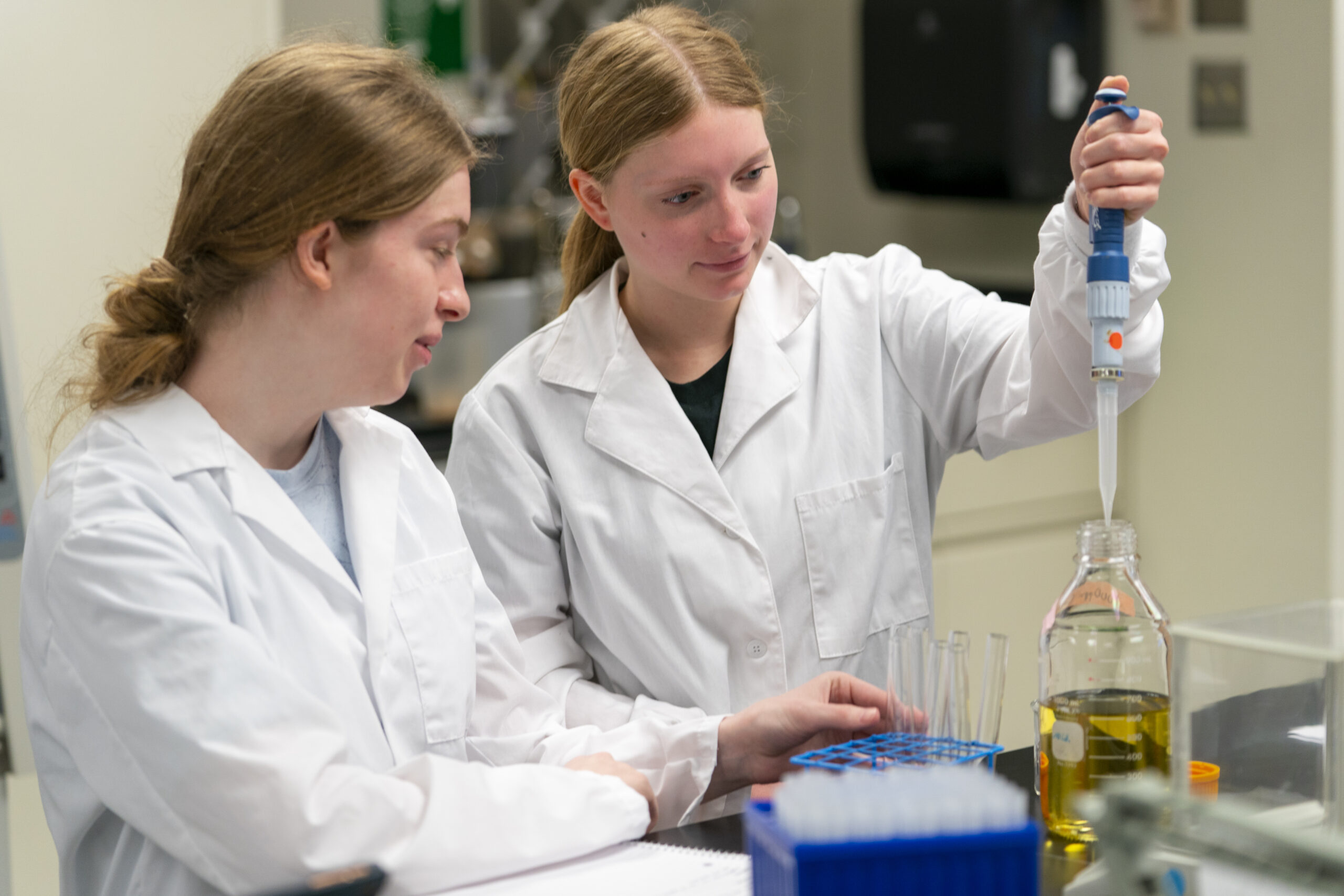 Students in lab wearing lab coats, one using a pipette to transfer liquid into a glass container