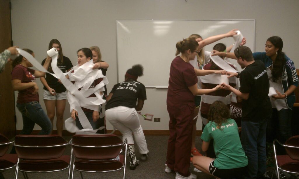 Students in a room wrapping two students with toilet paper