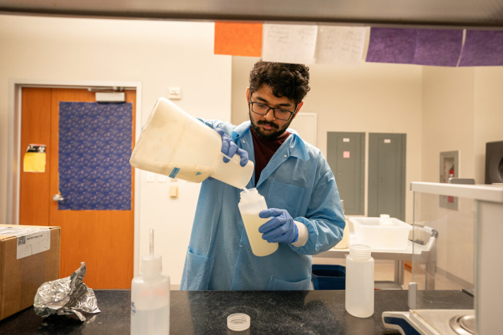 Man wearing glasses, gloves and lab coat pouring liquid from funnel-shapeed container into bottle.