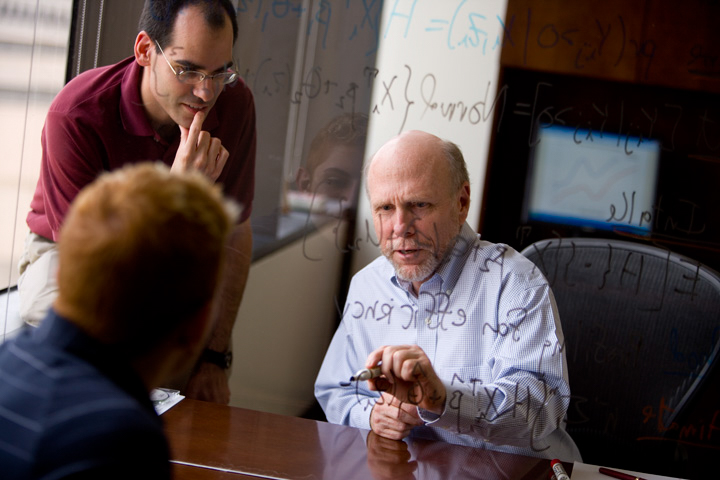 Professor writes on a clear board with an expo marker while talking to two men