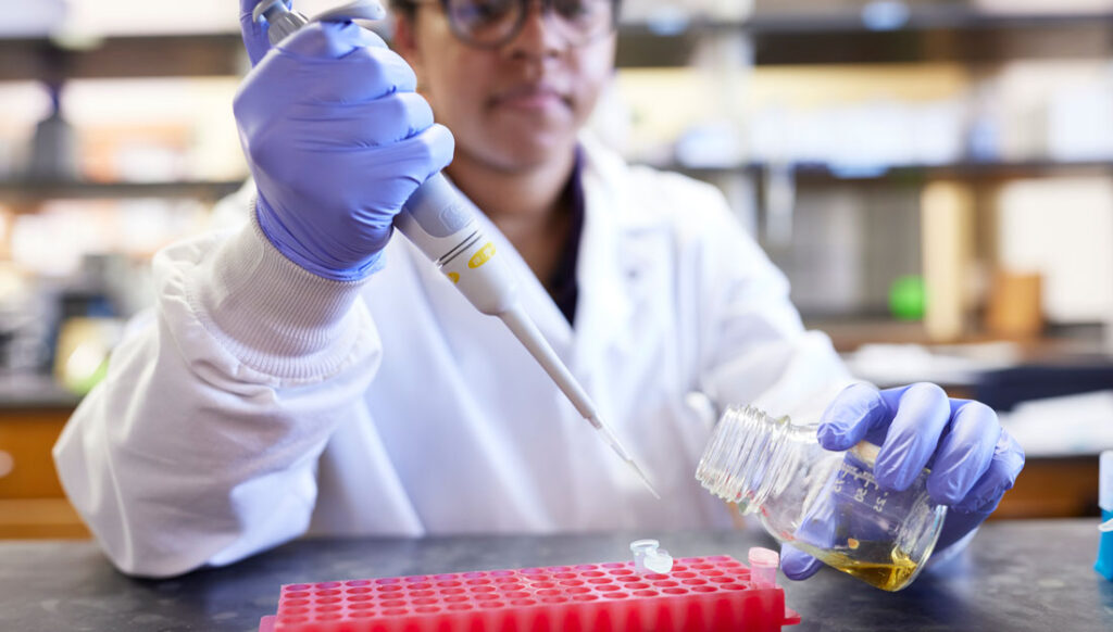 A student wearing a lab coat uses a pipette to transfer liquids from a vial to a bigger glass container