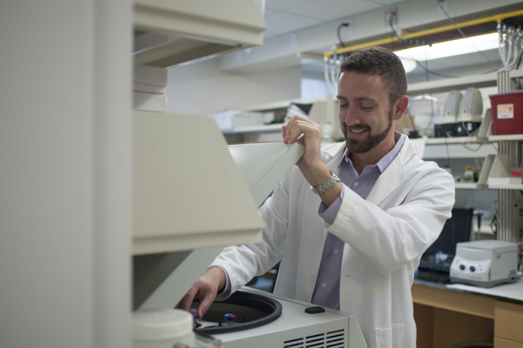 Student in a lab coat inside a lab opening a lid to a machine