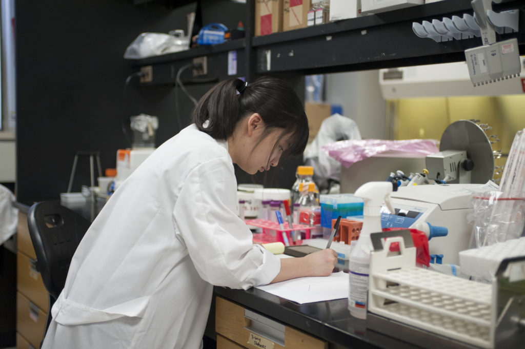 Student in a lab coat surrounded by lab supplies focussed on writing on a piece of paper